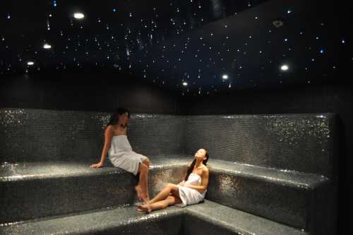 Two women in white towels relax on mosaic benches in a dimly lit spa with a starry ceiling.