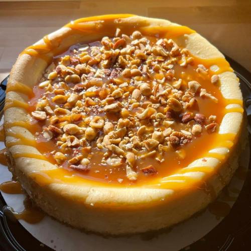 A creamy cheesecake topped with caramel sauce and chopped nuts on a black plate.