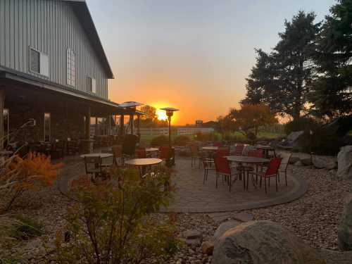 A serene outdoor patio with tables and chairs, framed by trees, as the sun sets in the background.