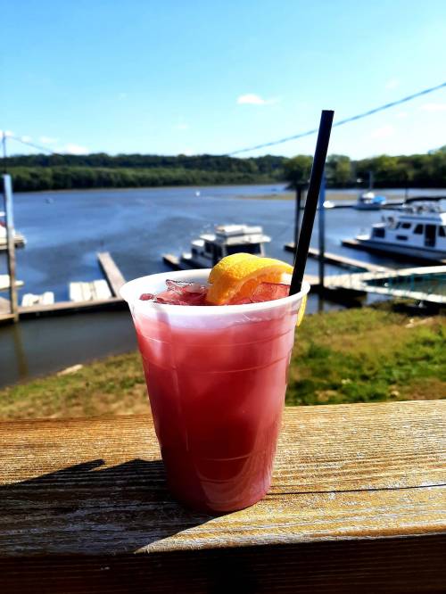 A refreshing drink with a lemon slice sits on a wooden railing, overlooking a marina and calm water under a blue sky.