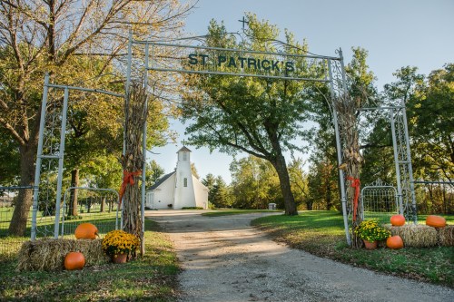 A decorative archway reading "St. Patrick's" leads to a white church, surrounded by trees, pumpkins, and hay bales.