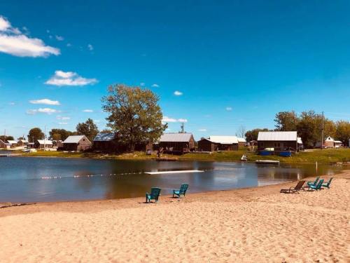 A sandy beach by a calm lake, with blue chairs and rustic buildings under a clear blue sky.