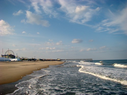 A sandy beach with gentle waves, a pier in the distance, and a ferris wheel under a partly cloudy sky.