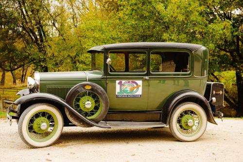 A vintage green car with a black roof parked on a gravel path, surrounded by trees.