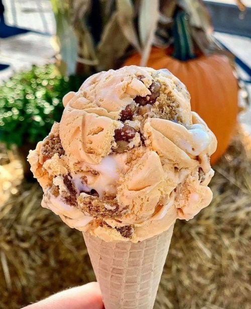 A close-up of a cone filled with creamy, swirled ice cream, featuring chunks and a warm autumn backdrop.