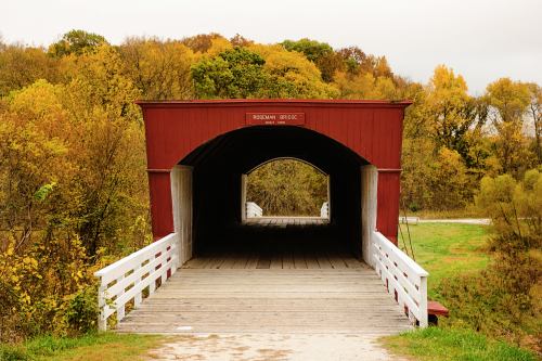 A red covered bridge with a wooden walkway, surrounded by trees displaying autumn colors.