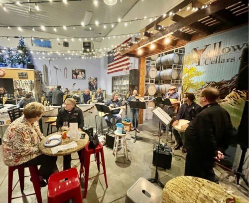 A lively indoor gathering with musicians performing, string lights overhead, and guests enjoying drinks at tables.