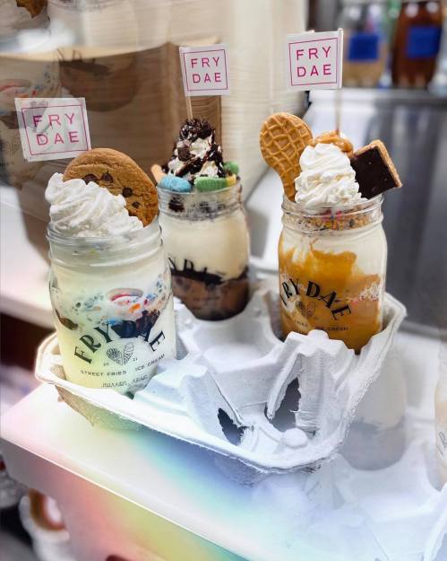 Three colorful ice cream desserts in jars, topped with whipped cream, cookies, and candy, displayed in a tray.