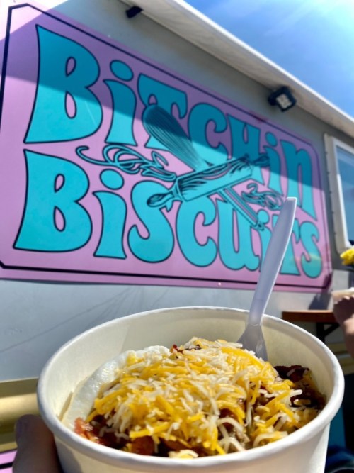 A bowl of cheesy food in front of a colorful food truck sign that reads "Bitchin Biscuits."