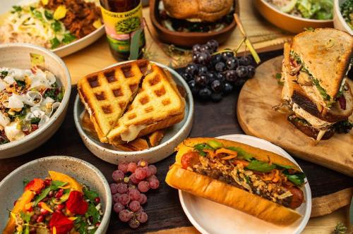 A colorful spread of various dishes, including sandwiches, salads, waffles, and hot dogs, with fresh grapes on the side.