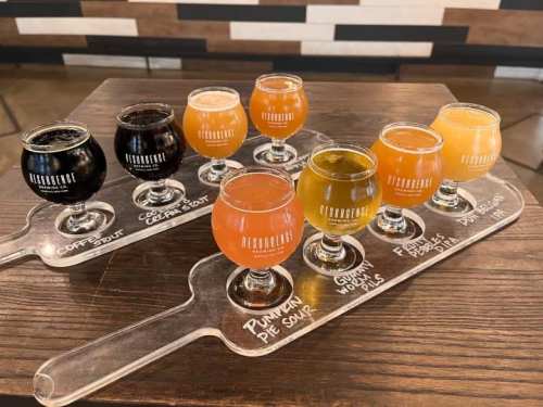 A flight of craft beers in small glasses on a wooden paddle, featuring various colors and styles.