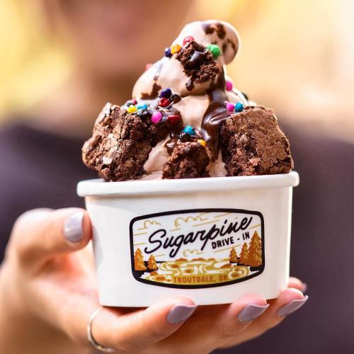 A hand holds a cup of chocolate soft serve topped with brownie pieces and colorful sprinkles.