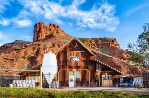A rustic brewery building with a "BEER" sign, surrounded by barrels and set against a rocky hillside and blue sky.