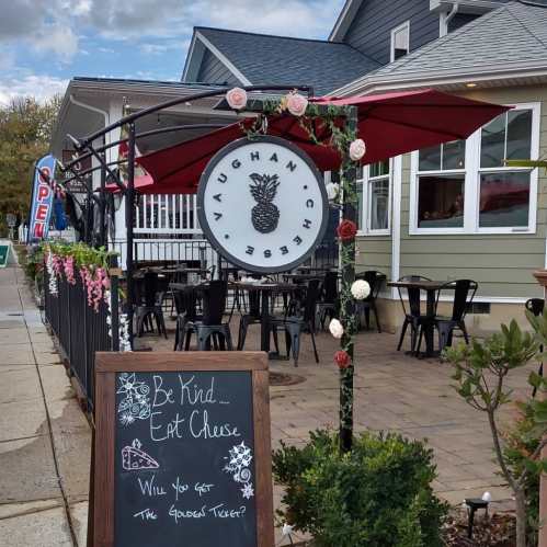 A charming cheese shop entrance with a sign, outdoor seating, and decorative flowers. A chalkboard promotes kindness and cheese.