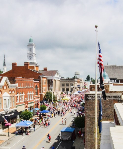 A bustling street festival with crowds, flags, and historic buildings under a cloudy sky.
