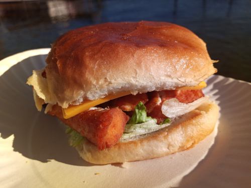 A crispy chicken sandwich with lettuce and cheese on a soft bun, served on a paper plate by a body of water.