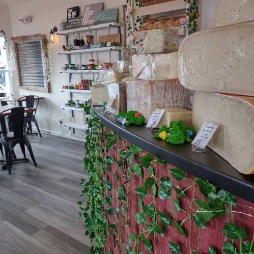 A cozy cheese shop interior featuring various cheese blocks on a counter, surrounded by greenery and shelves of products.