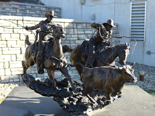 Bronze sculpture of two cowboys on horseback roping a steer, capturing a dynamic rodeo scene.