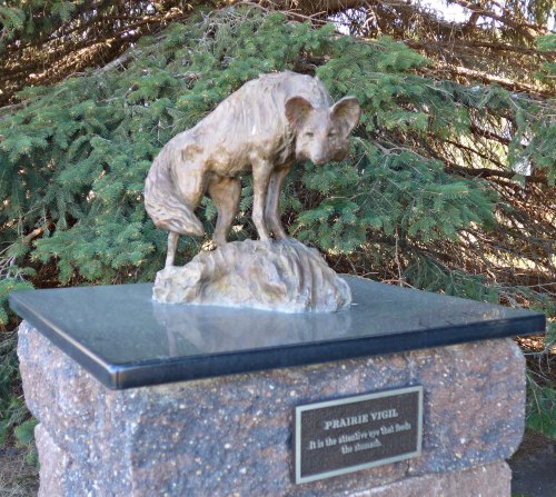 A bronze sculpture of a fox standing on a rock, titled "Prairie Vigil," on a stone pedestal surrounded by greenery.
