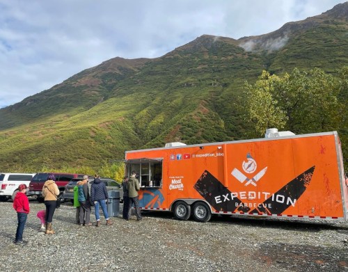 A food truck named "Expedition Barbecue" parked in a scenic area, with people lined up to order. Mountains in the background.