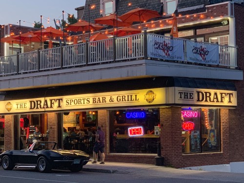 A sports bar and grill named "The Draft" with outdoor seating, neon signs, and a classic car parked outside.