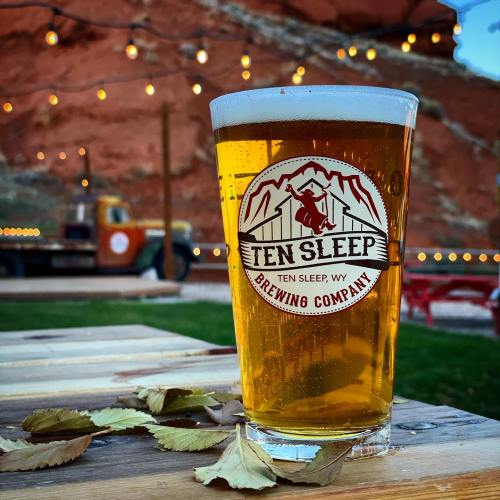 A glass of beer sits on a wooden table, with a rustic backdrop of red rock and string lights in the evening.
