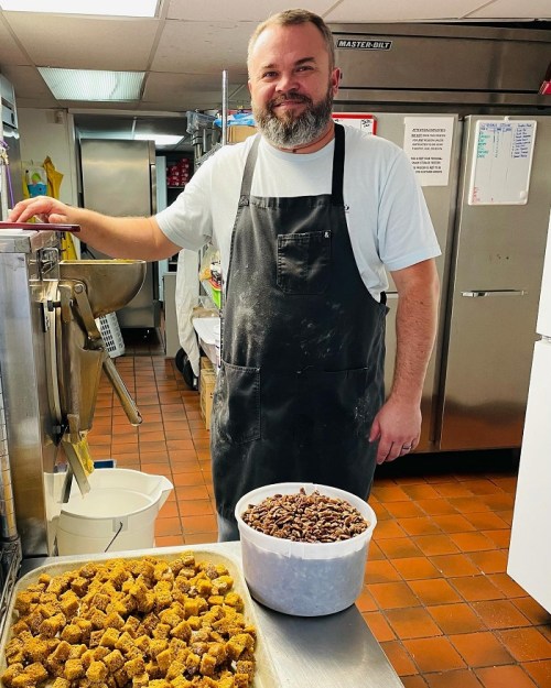 A smiling man in an apron stands in a kitchen next to trays of honeycomb and a bucket of pecans.