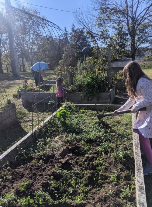 A girl with long hair digs in a garden while two people work in the background, surrounded by plants and trees.
