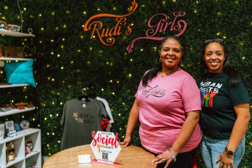 Two women smile at a table in a vibrant shop decorated with greenery and lights, showcasing merchandise and a sign.