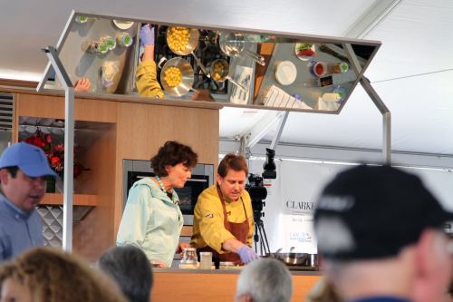 A chef and a woman prepare food on stage, with a mirrored surface reflecting their actions and audience in a tent.