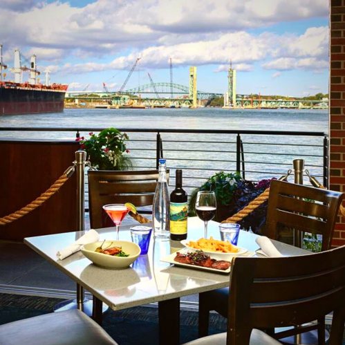 A scenic riverside dining table with a steak, salad, and drinks, overlooking a bridge and ships in the background.