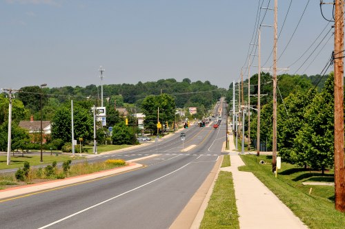 A wide road stretches into the distance, lined with trees and power lines under a clear blue sky.