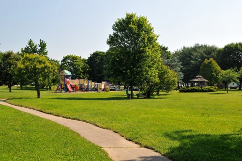 A sunny park with green grass, trees, a playground in the background, and a winding path leading through the area.