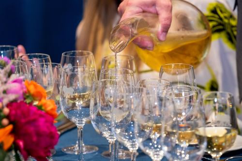 A person pours a golden liquid into a row of clear glasses, surrounded by colorful flowers on a table.