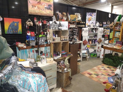 A vibrant market stall filled with handmade crafts, jewelry, and colorful decor items, set in a spacious indoor venue.