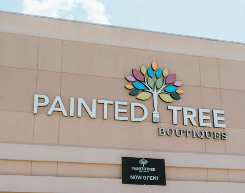 Sign for Painted Tree Boutiques featuring a colorful tree logo and a banner stating "NOW OPEN!"