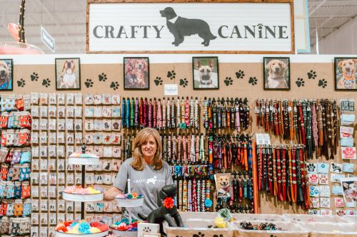 A woman stands behind a display of colorful dog accessories at a pet shop called "Crafty Canine."