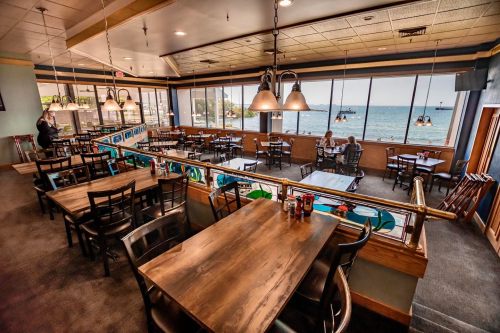 A spacious restaurant with wooden tables and chairs, overlooking a scenic view of the water and boats outside.
