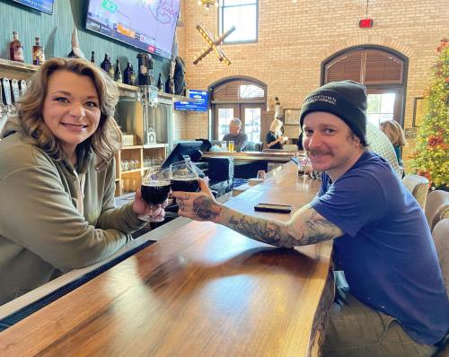 Two people at a bar clink glasses of dark drinks, smiling and enjoying their time together.