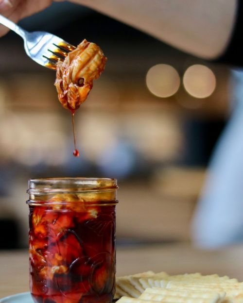 A fork lifts a piece of food from a jar filled with sauce, next to a plate of crackers on a wooden table.