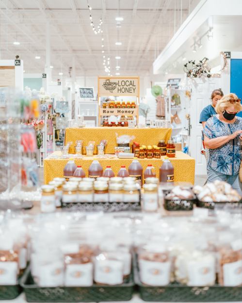 A market scene featuring a display of local honey products, with shoppers browsing in the background.