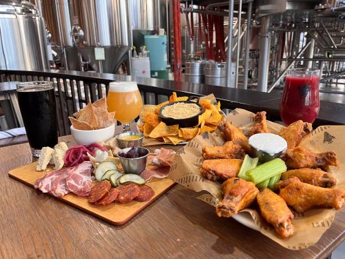 A wooden table with a platter of chicken wings, dips, and a charcuterie board, alongside various craft beers.