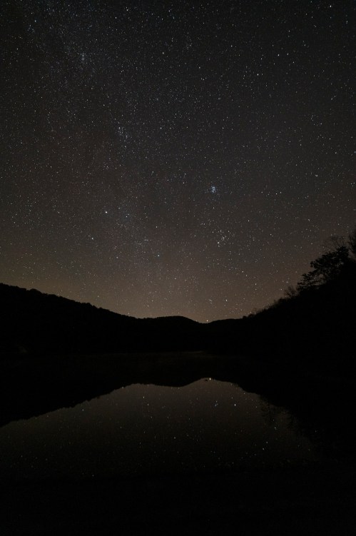 A starry night sky reflected in a calm lake, surrounded by dark silhouettes of mountains.
