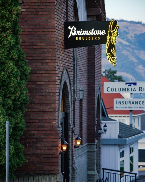 A neon sign for "Brimstone Boulders" hangs above a brick building, with trees and mountains in the background.