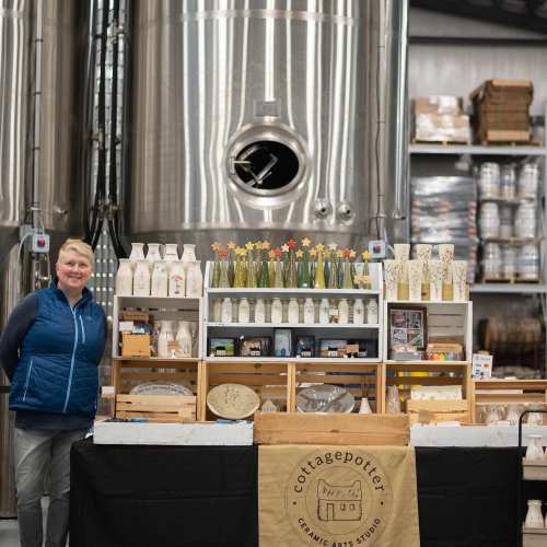 A smiling woman stands beside a display of pottery in a studio with large brewing tanks in the background.