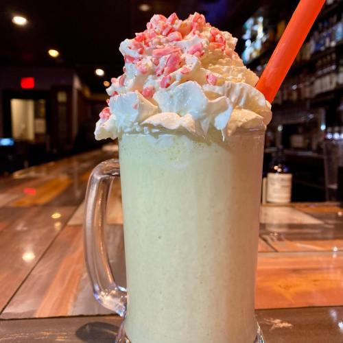 A tall glass of creamy milkshake topped with whipped cream and crushed peppermint, served with an orange straw.