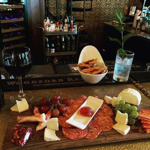 A charcuterie board with meats, cheeses, grapes, and a glass of red wine on a bar counter.