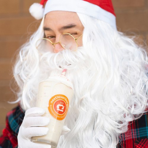 A person dressed as Santa Claus enjoys a festive milkshake, wearing glasses and a red hat with a white beard.