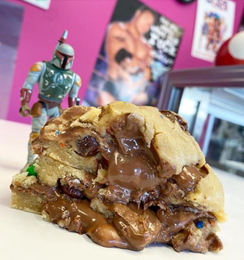 A gooey chocolate chip cookie stack with colorful sprinkles, next to a Boba Fett action figure against a pink background.
