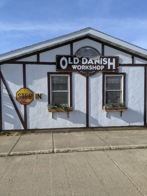 A quaint building with a sign reading "Old Danish Workshop" and a "Stop In" sign, featuring flower boxes under the windows.